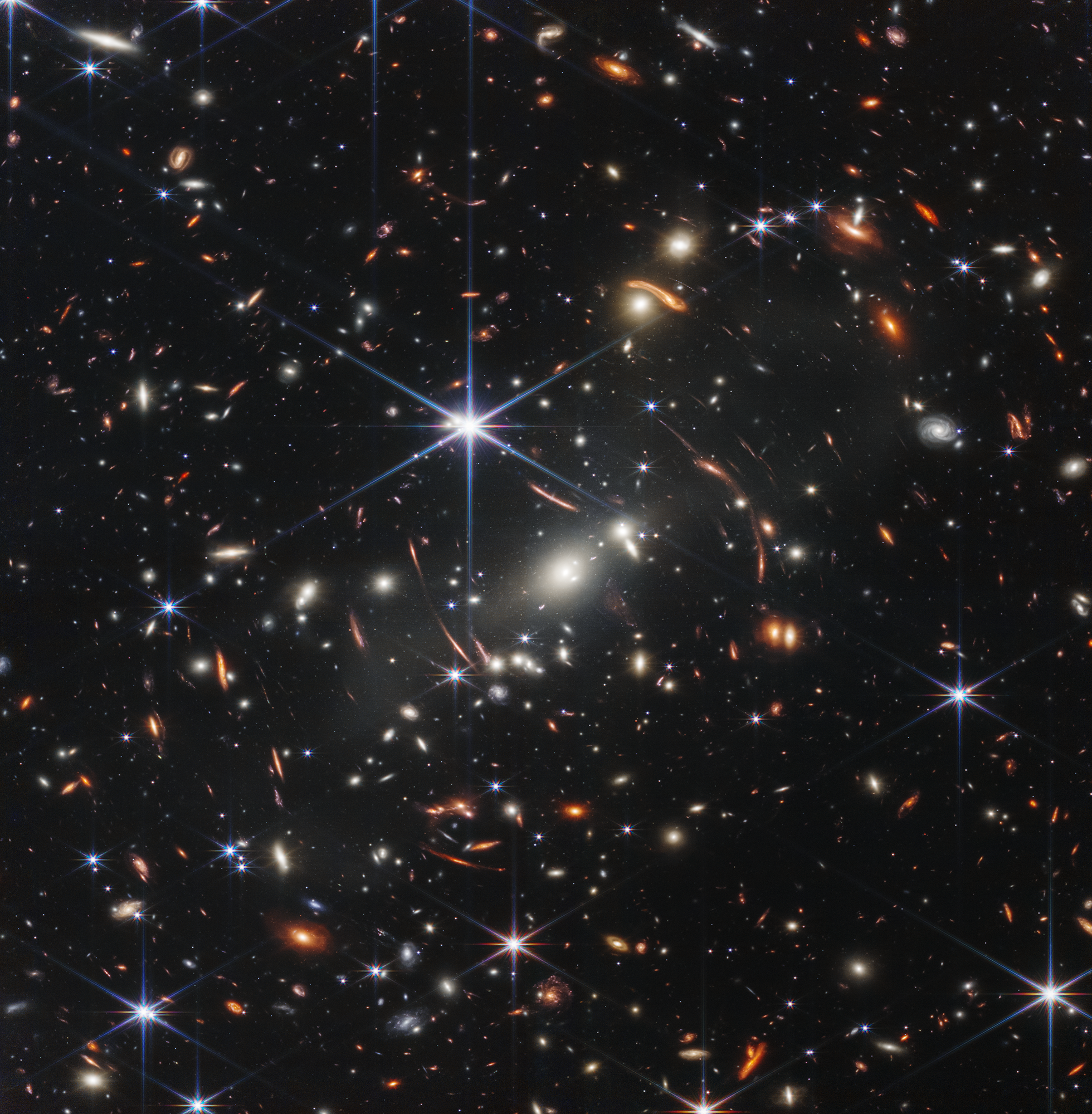 Thousands of small galaxies appear across this view. Their colors vary. Some are shades of orange, while others are white. Most appear as fuzzy ovals, but a few have distinct spiral arms. In front of the galaxies are several foreground stars. Most appear blue, and the bright stars have diffraction spikes, forming an eight-pointed star shape. There are also many thin, long, orange arcs that curve around the center of the image.