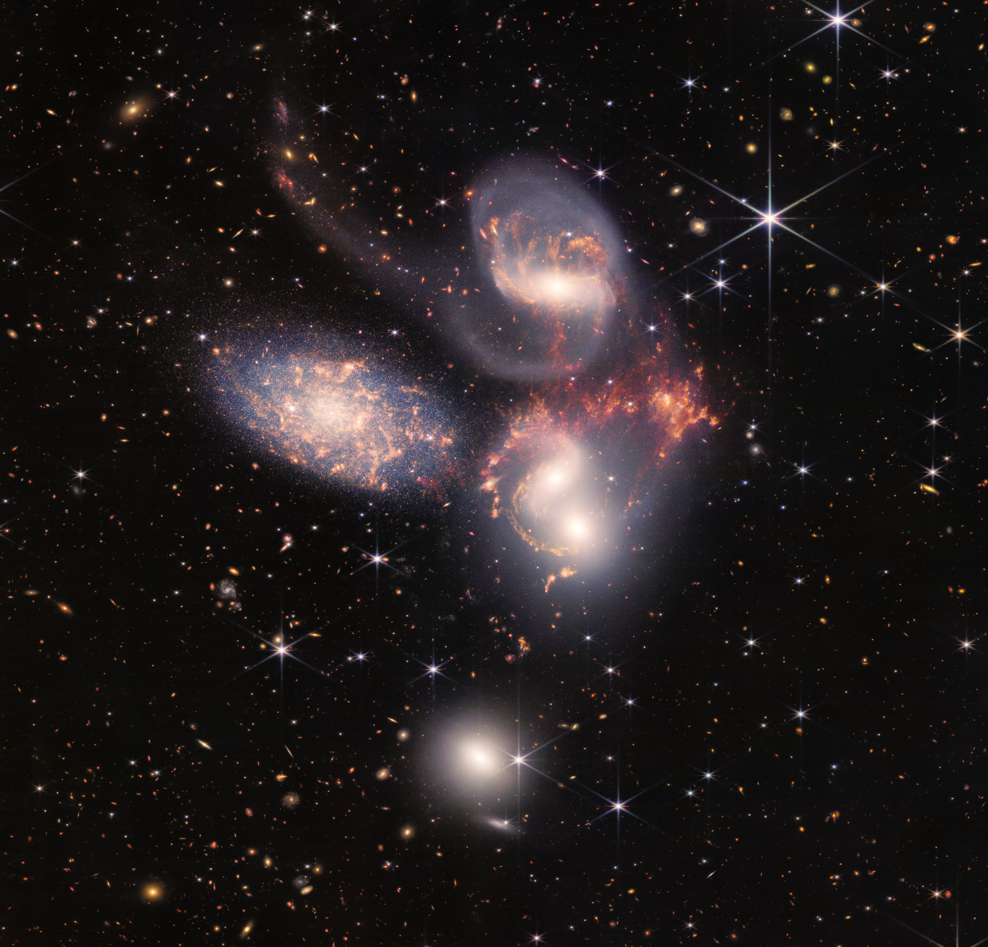 Image of a group of five galaxies that appear close to each other in the sky: two in the middle, one toward the top, one to the upper left, and one toward the bottom. Four of the five appear to be touching. One is somewhat separated. In the image, the galaxies are large relative to the hundreds of much smaller (more distant) galaxies in the background. All five galaxies have bright white cores. Each has a slightly different size, shape, structure, and coloring. Scattered across the image, in front of the galaxies are number of foreground stars with diffraction spikes: bright white points, each with eight bright lines radiating out from the center.